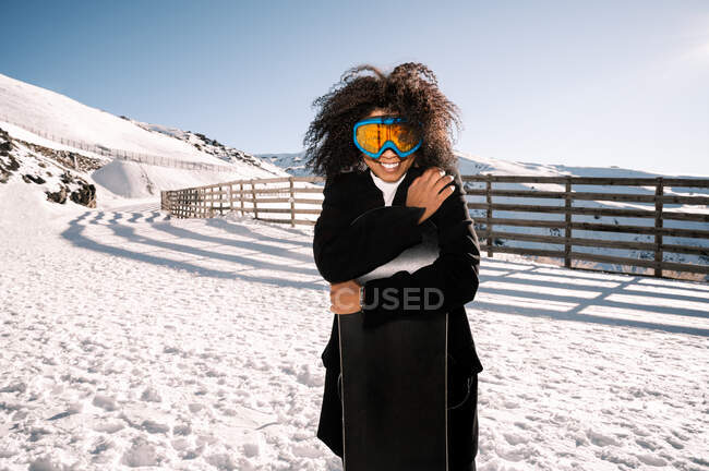 Happy ethnic sportswoman in stylish wear and protective glasses embracing snowboard on snowy mount in sunlight — Stock Photo