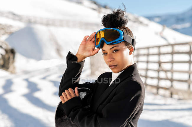 Jolie athlète afro-américaine en tenue tendance et lunettes de protection debout avec snowboard regardant la caméra en hiver — Photo de stock