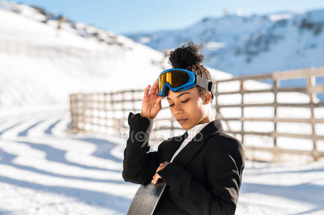 Jolie athlète afro-américaine en tenue tendance et lunettes de protection debout avec snowboard regardant vers le bas en hiver — Photo de stock