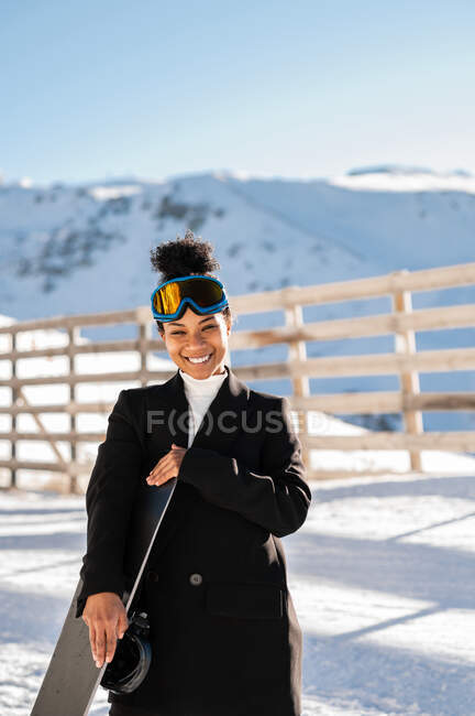 Mujer deportista étnica feliz en ropa elegante y gafas protectoras que llevan snowboard en montura nevada bajo la luz del sol mirando a la cámara - foto de stock