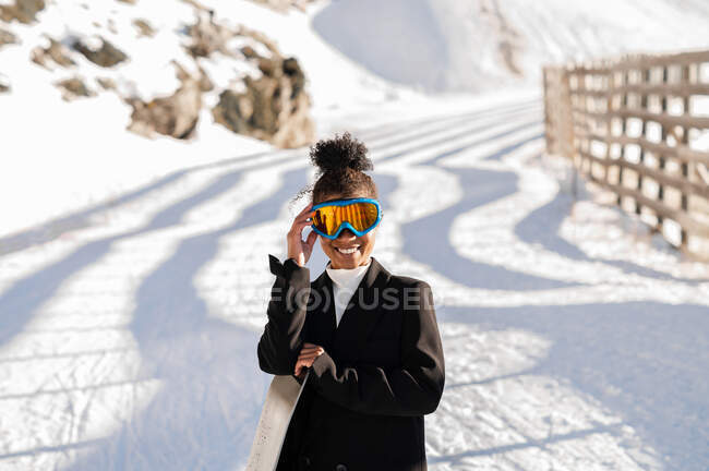 Desportista étnica feliz no desgaste elegante e óculos de proteção carregando snowboard no monte nevado na luz do sol olhando para a câmera — Fotografia de Stock