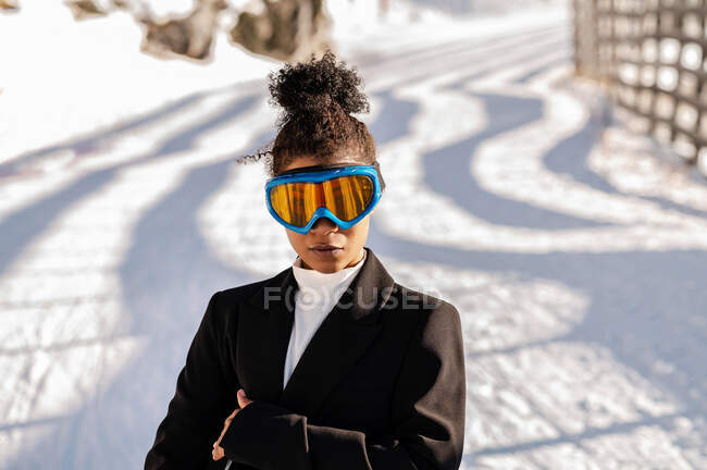 Stylish young ethnic female athlete in protective glasses looking at camera on snowy roadway in sunlight — Stock Photo