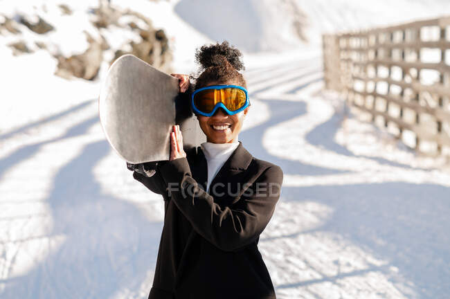 Счастливая этническая спортсменка в стильной одежде и защитных очках, несущая сноуборд на снежной горе под солнечным светом, смотрящая в камеру — стоковое фото