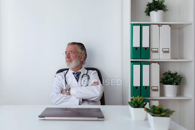 Dreamy anciano médico en gafas con estetoscopio mirando a la mesa con la tableta en el hospital - foto de stock