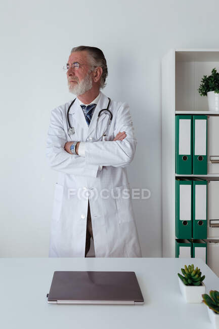 Старший врач-мужчина с седой бородой и сложенными руками в халате со стетоскопом, смотрящим в клинике — стоковое фото
