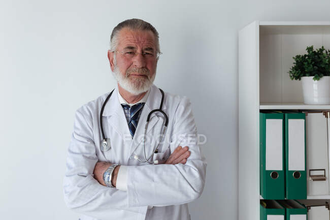 Старший врач-мужчина с седой бородой и сложенными руками в халате со стетоскопом, смотрящий в камеру в клинике — стоковое фото