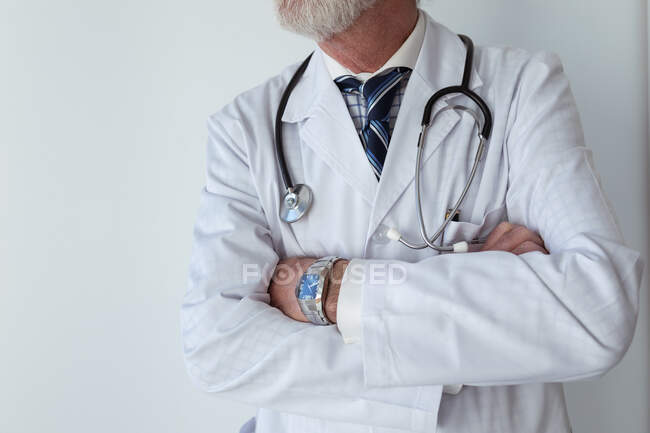 Crop senior medico maschio con barba grigia e braccia piegate in accappatoio con stetoscopio in piedi in clinica — Foto stock