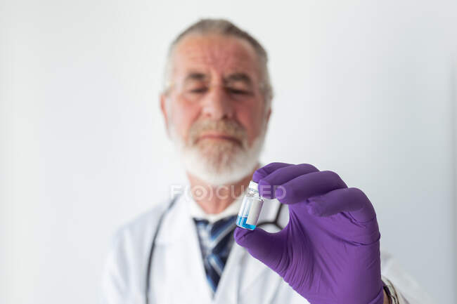 Médico barbudo mayor en guantes uniformes y desechables demostrando una pequeña botella con sustancia líquida azul sobre fondo blanco - foto de stock
