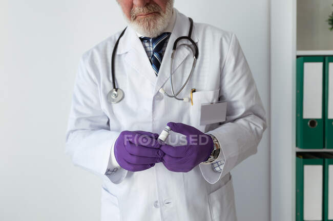 Crop senior bearded male doctor in uniform and disposable gloves demonstrating small bottle with blue liquid substance on white background — Stock Photo