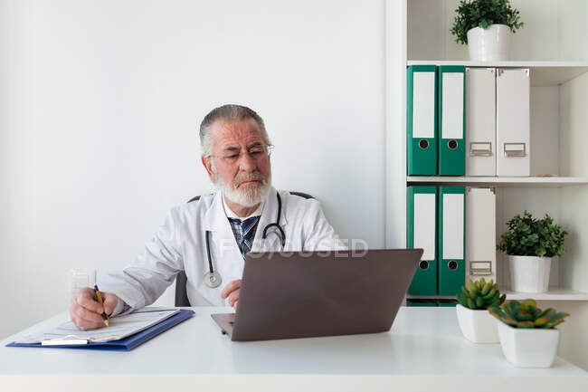 Médecin principal en uniforme écrivant des notes de service sur papier pendant la consultation en ligne sur l'ordinateur portable à l'hôpital — Photo de stock