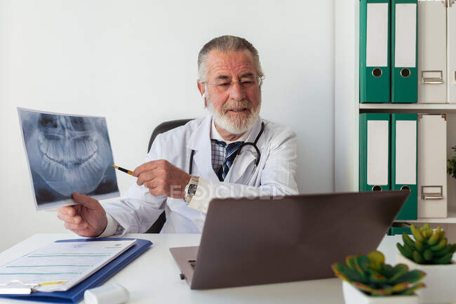Elderly male orthodontist showing X ray image of teeth during video chat on netbook at table in hospital — Stock Photo