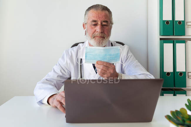 Elderly bearded male physician in wireless earbud demonstrating disposable mask against netbook during online health consultation in hospital — Stock Photo