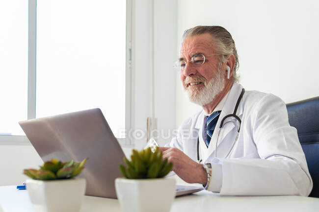 Elderly male doctor in earbud showing medications while talking on video chat against netbook during online health consultation in hospital — Stock Photo