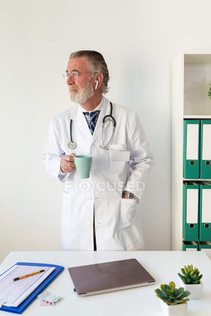 Senior medico di sesso maschile in uniforme con stetoscopio con bevanda calda guardando lontano in ospedale alla luce del giorno — Foto stock