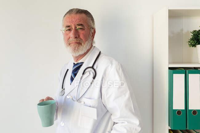 Médico do sexo masculino em uniforme com estetoscópio com bebida quente olhando para a câmera no hospital à luz do dia — Fotografia de Stock