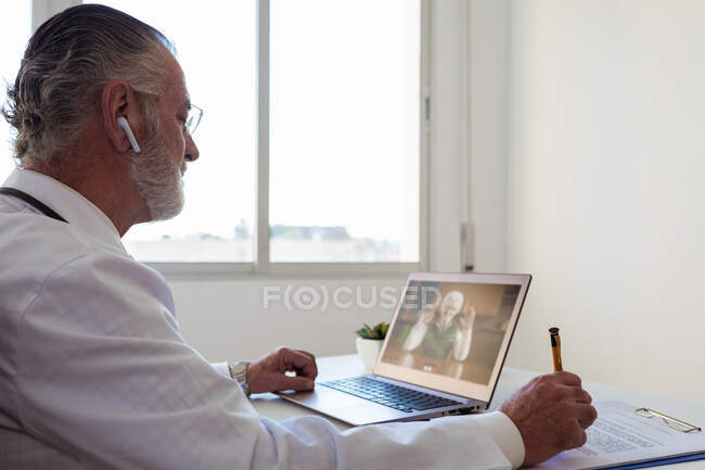 Side view of elderly male doctor in wireless earbud speaking with woman on video call on netbook in clinic — Stock Photo