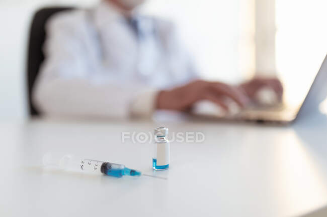 Crop médico anónimo trabajando en netbook en la mesa con inyector y botella pequeña con sustancia azul en la clínica - foto de stock