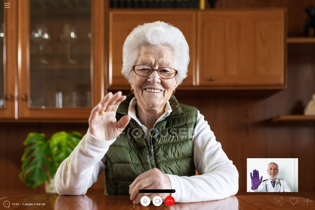 Friendly elderly female showing greeting gesture against tablet while video chatting in house — Stock Photo
