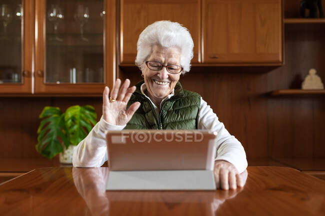 Friendly elderly female showing greeting gesture against tablet while video chatting in house — Stock Photo