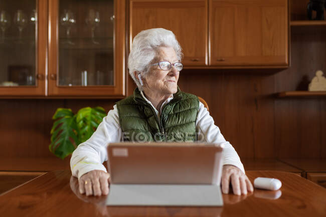 Friendly elderly female in wireless earbuds looking away while video chatting in house — Stock Photo