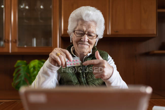 Mujer anciana en auricular mostrando medicamentos mientras habla en video chat contra netbook durante la consulta de salud en línea en el hogar - foto de stock