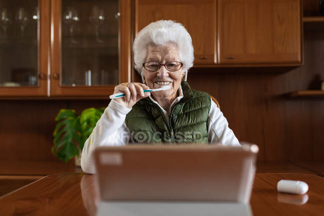 Elderly woman with gray hair in wireless earbuds with toothbrush against tablet during online health consultation in house — Stock Photo