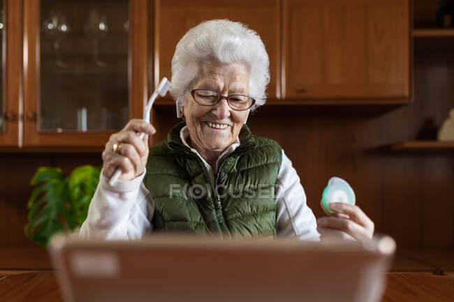 Mujer anciana con pelo gris en auriculares inalámbricos con cepillo de dientes y hilo dental contra la tableta durante la consulta de salud en línea en casa - foto de stock