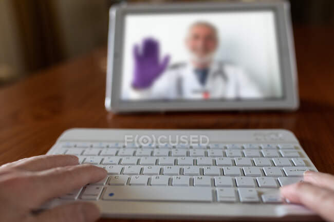 Crop anonymous patient with keyboard against tablet with doctor on screen during video call in house — Stock Photo