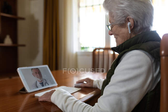 Side view of crop elderly female patient in TWS earbuds speaking with doctor on tablet during video call at home — Stock Photo