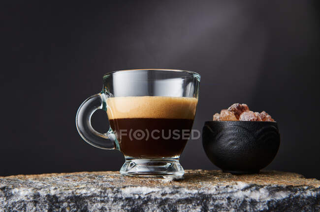 Transparent glass of strong black coffee with foam near bowl of brown sugar cubes on black background — Stock Photo