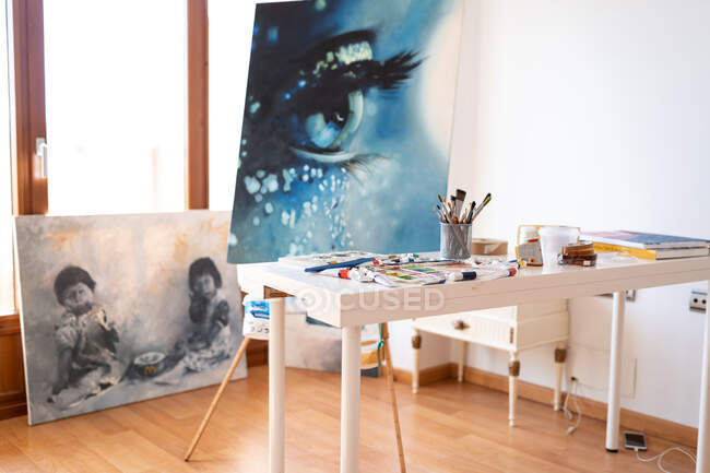 Art studio interior with collection of painted pictures and paint brushes in glass on table in daylight — Stock Photo