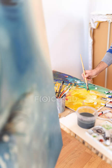 Crop unrecognizable male painter using professional brush during painting process on carton sheet near art tools in workroom — Stock Photo