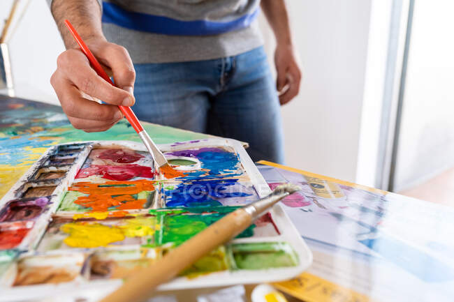 Анонимный мужчина-художник, смешивающий краски с кистью, используя палитру акварели во время работы в художественной студии — стоковое фото