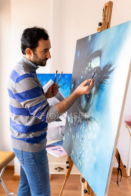 Vue de côté du jeune peintre ethnique barbu en tenue décontractée peinture avec pinceau sur toile en atelier d'art — Photo de stock