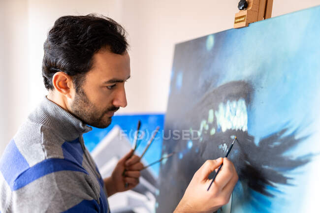 Side view of young bearded ethnic painter in casual wear painting with brush on canvas in art studio — Fotografia de Stock