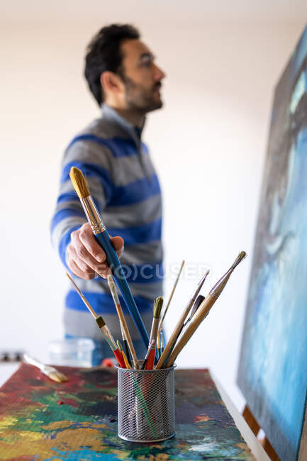 Blurred anonymous male painter taking paint brush from glass near painting representing eye in workroom - foto de stock