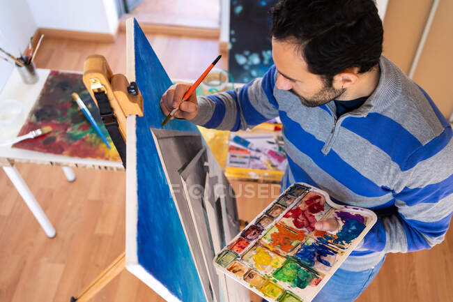 From above young bearded ethnic painter in casual wear painting with brush on canvas in art studio — Foto stock