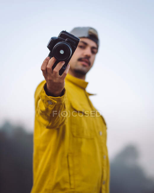 Side view content young male photographer in yellow denim jacket taking photo on photo camera and looking at camera while standing on foggy blurred nature background - foto de stock