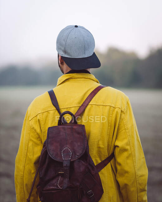 Back view anonymous male traveler in yellow denim jacket and cap standing on vast lawn on foggy gloomy day - foto de stock