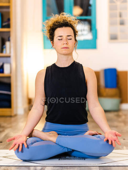 Mujer tranquila en ropa deportiva sentada en la esterilla y pose de loto mientras practica yoga en un estudio de luz - foto de stock