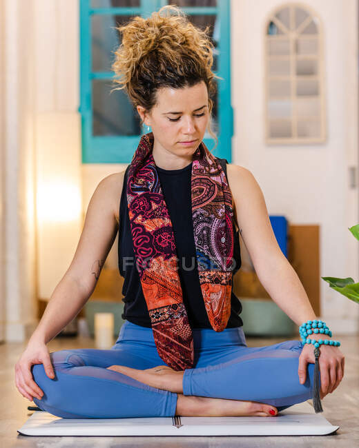 Calm female in activewear sitting on mat on Half Lotus meditating with mala beads on arm in light studio — Stock Photo