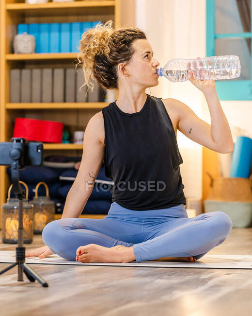 Спокійна жінка з кучерявим волоссям у спортивному одязі, що сидить на підлозі та п'є пляшку води під час перерви в онлайн-сесії йоги в студії — стокове фото