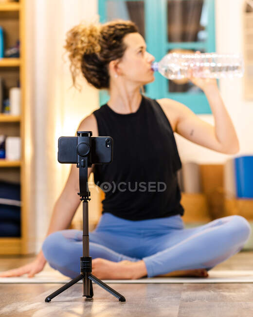 Спокойная женщина с кудрявыми волосами в спортивной одежде сидит на полу и пьет бутылку воды во время перерыва в онлайн-сессии йоги в студии — стоковое фото