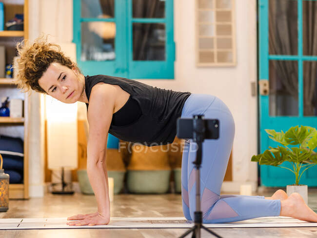 Vista lateral da jovem mulher concentrada em sportswear fazendo pose de mesa e olhando para a tela do smartphone enquanto pratica ioga em aula on-line em estúdio — Fotografia de Stock