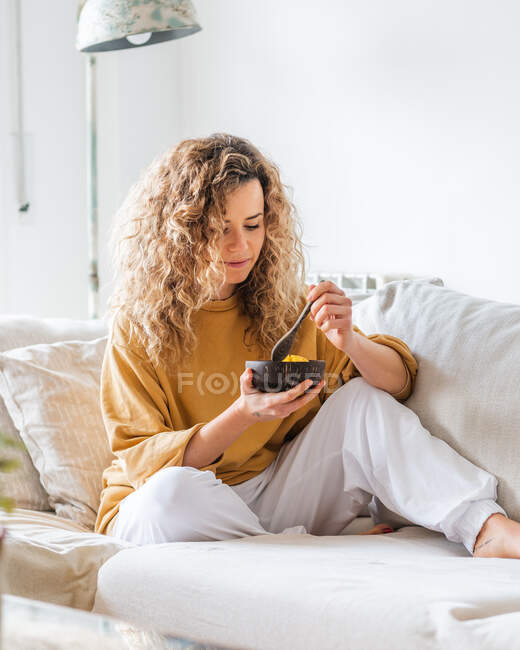 Jovem loira com cabelos cacheados em roupas casuais sentada no sofá confortável enquanto come comida saudável na sala de luz — Fotografia de Stock