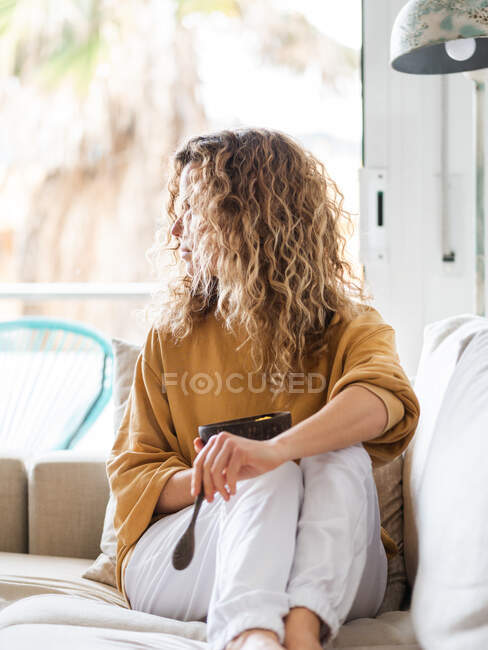 Junge blonde Frau mit lockigem Haar in lässiger Kleidung sitzt auf einem bequemen Sofa, während sie in einem hellen Raum gesunde Nahrung zu sich nimmt — Stockfoto