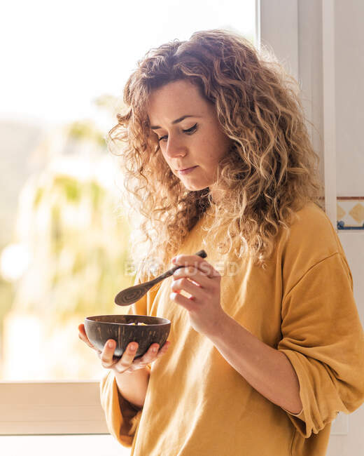 Pensive female with curly hair in casual clothes standing with wooden spoon and bowl with food in light room against window in daytime — Stock Photo