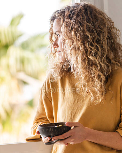 Pensive female with curly hair in casual clothes standing with wooden spoon and bowl with food in light room against window in daytime — Stock Photo