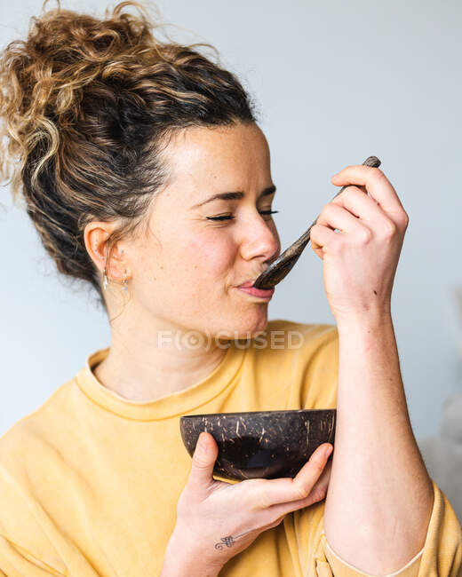 Молода блондинка весела жінка в повсякденному одязі їсть здорову їжу з натуральної кокосової миски в світлій кімнаті — стокове фото