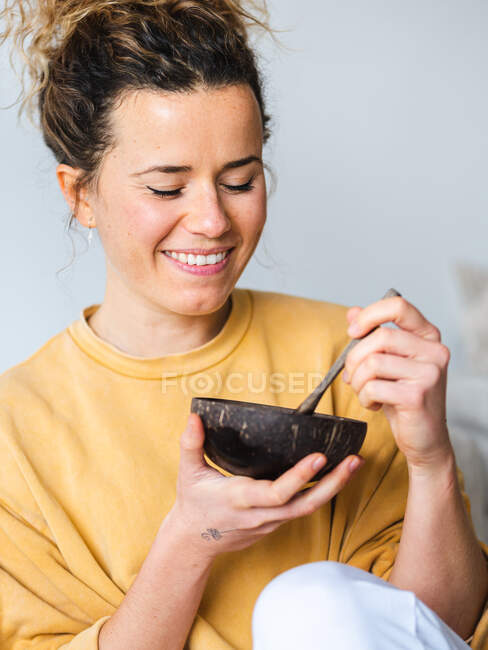 Giovane donna bionda allegra in abiti casual mangiare cibo sano dalla ciotola di cocco naturale in camera luce — Foto stock